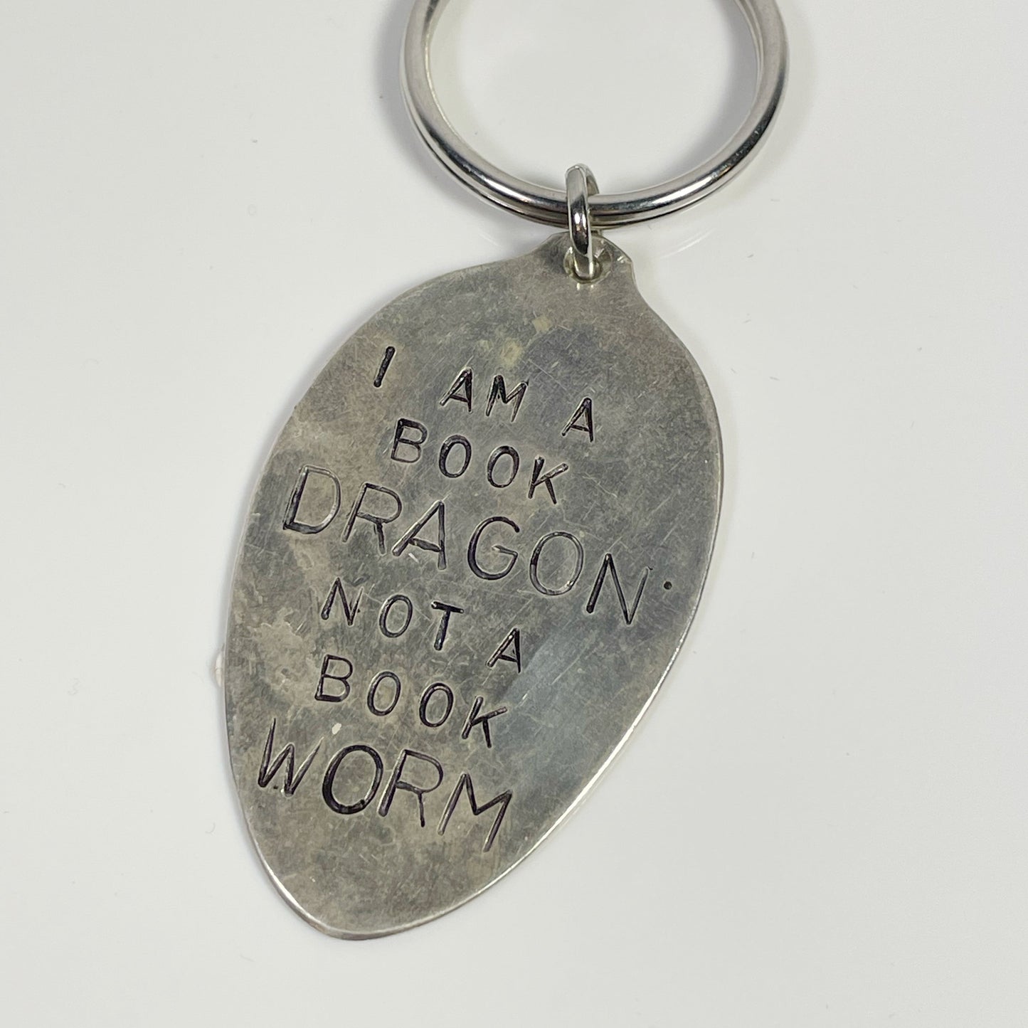 A silver-colored keychain with a stamped message that reads 'I am a BOOK DRAGON not a BOOK WORM'.