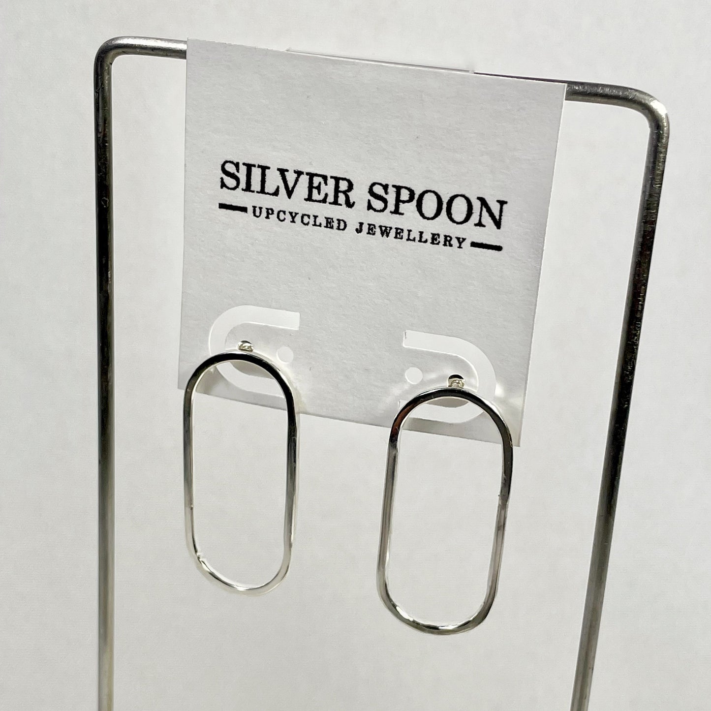 A pair of silver oval earrings displayed on a silver spoon stand, with the brand name 'Silver Spoon' visible on a tag.