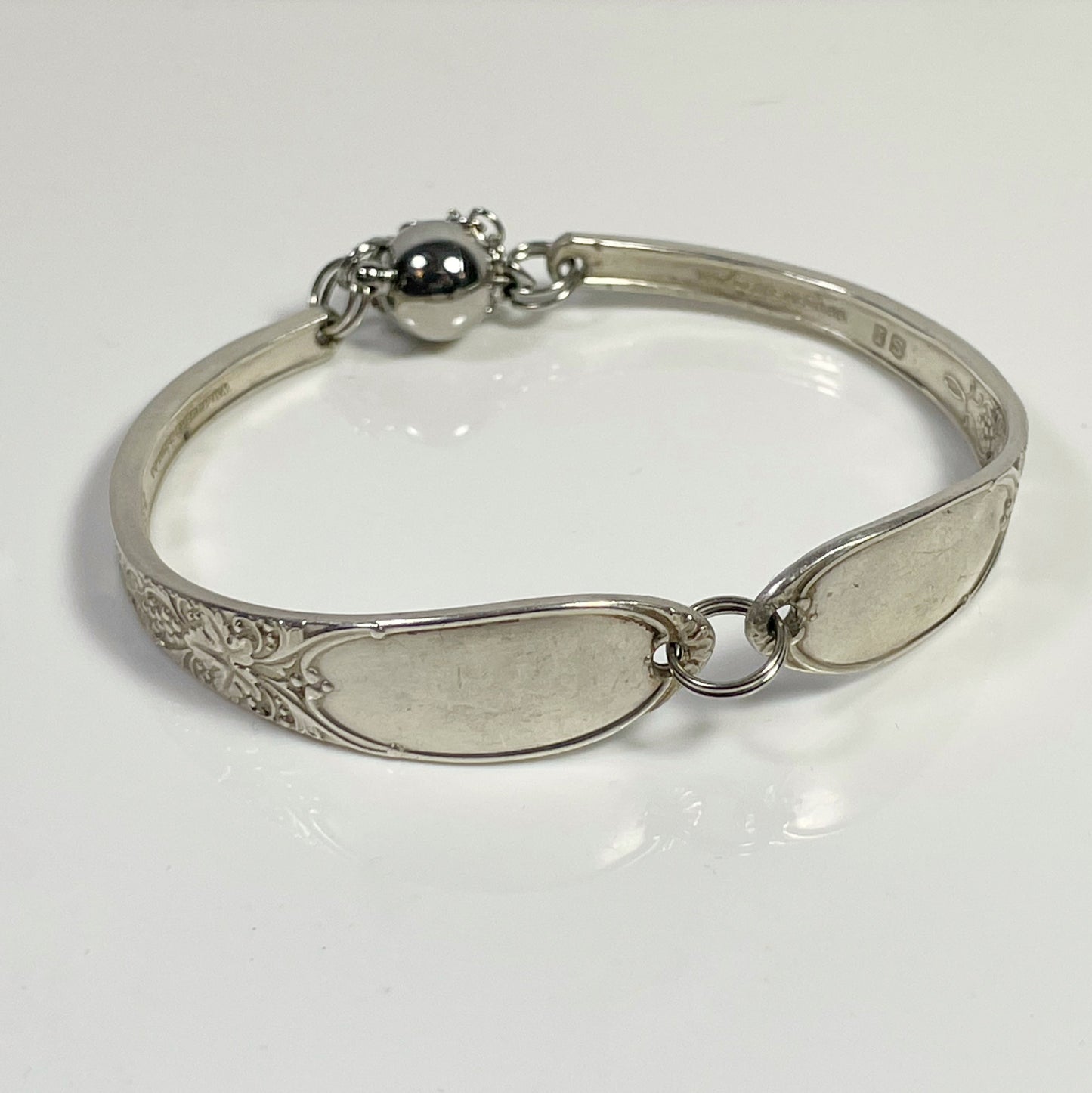 A silver bracelet made from two spoon handles, featuring a clasp and a decorative design, displayed against a white background.