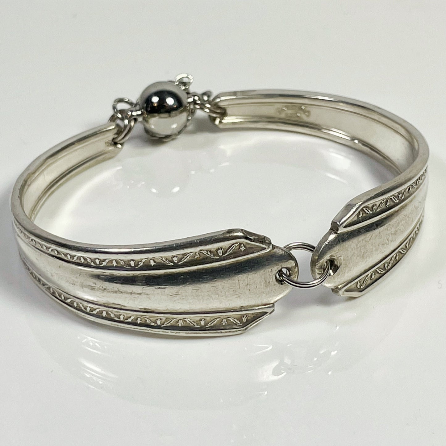 A silver spoon handle bracelet with a split jump ring and decorative patterns, featuring a strong magnetic clasp with safety chain.