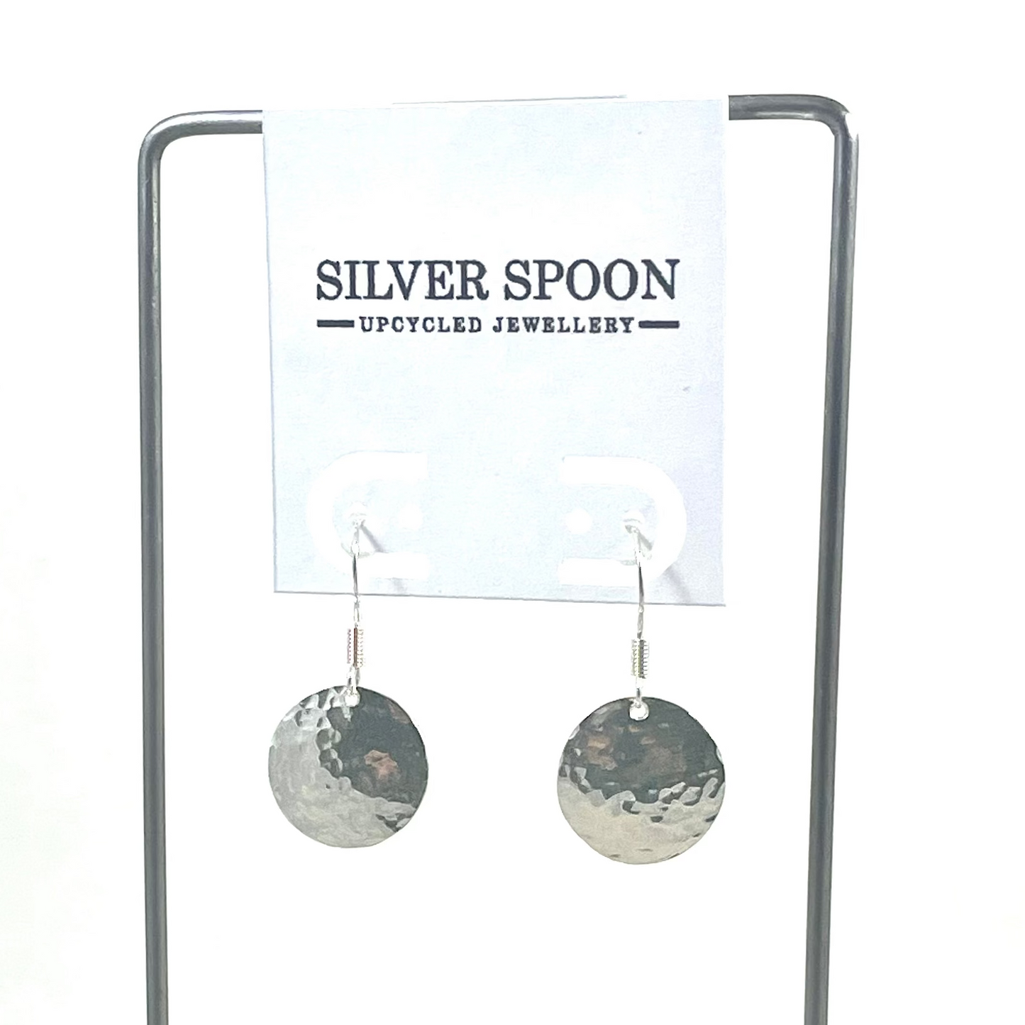 A pair of sterling silver hammered disk earrings displayed on a wooden stand with a branded card above.