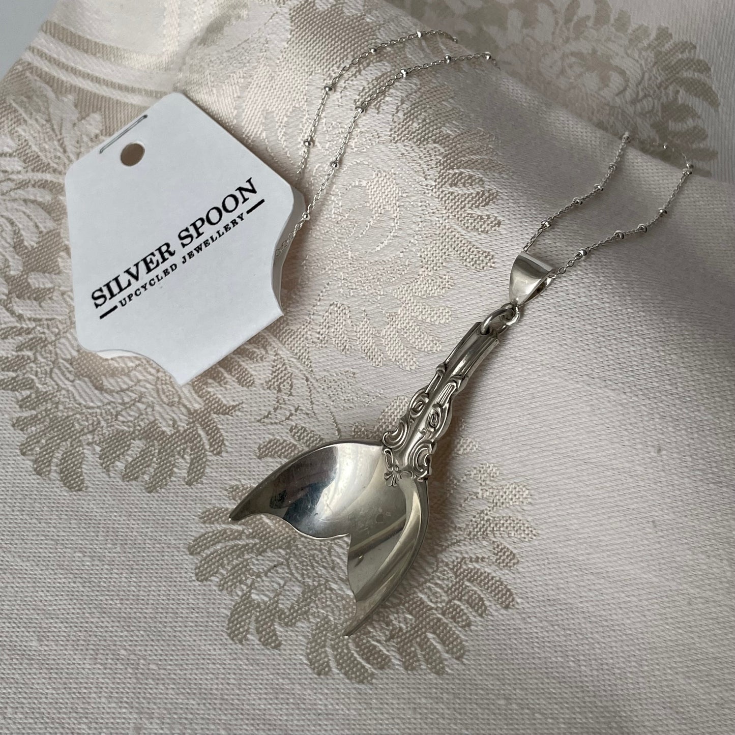 Silver spoon with a decorative handle on a textured fabric background, featuring a 'Silver Spoon' tag.